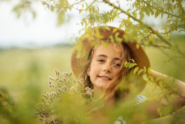 Kleines Mädchen mit lockigem Haar in einem Hut und Blumen steht in einem langen Kleid