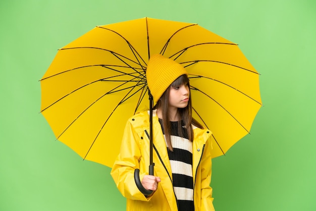 Kleines Mädchen mit regenfestem Mantel und Regenschirm über isoliertem Chroma-Key-Hintergrund mit Blick zur Seite