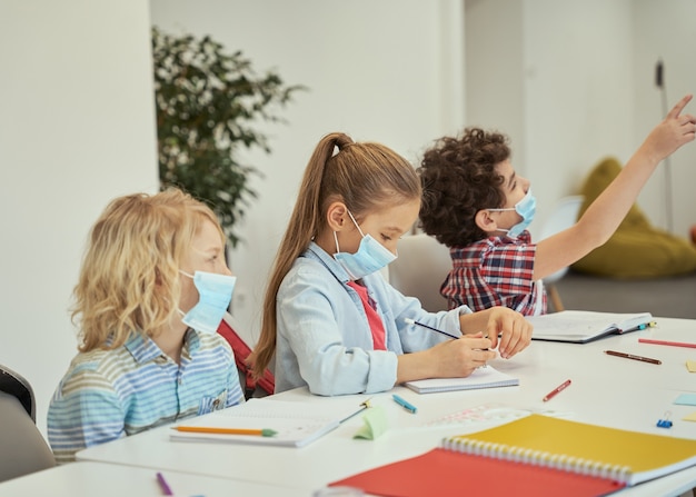 Kleines Mädchen mit Schutzmaske, das ihr Notizbuch anschaut Grundschulkinder hören zu