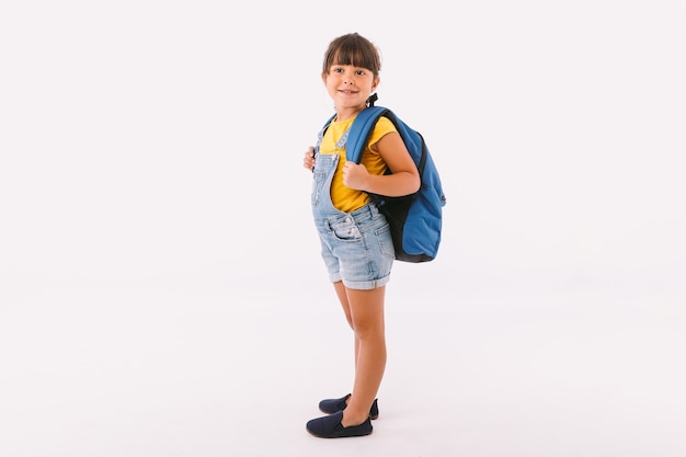 Kleines Mädchen mit schwarzen Haaren, gekleidet in einen Denim-Overall und ein blaues T-Shirt, mit einem Rucksack, der bereit ist, wieder zur Schule zu gehen, auf ihrer Seite, auf weißem Hintergrund