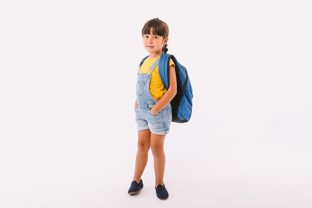 Kleines Mädchen mit schwarzen Haaren, gekleidet in einer blauen Latzhose und einem T-Shirt, mit einem Rucksack, der für den Schulanfang bereit ist, auf weißem Hintergrund