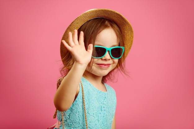 Kleines Mädchen mit Sommerhut und Sonnenbrille winkt dir zu und drückt Freude und Glück aus, die auf Rosa stehen