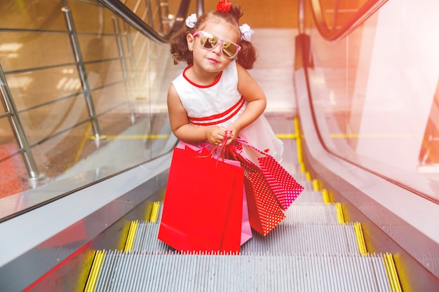 Kleines Mädchen mit Sonnenbrille auf der Rolltreppe im Einkaufszentrum mit Einkäufen