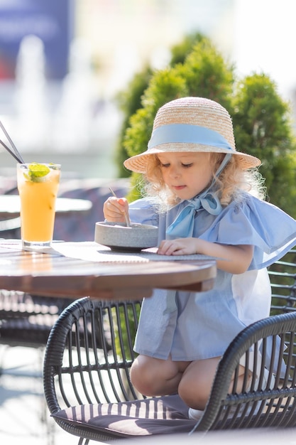 Kleines Mädchen mit Strohhut und blauem Kleid frühstückt im Freien