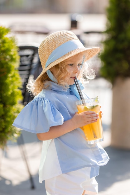 Kleines Mädchen mit Strohhut und blauem Kleid trinkt Saft aus einem Glas im Freien