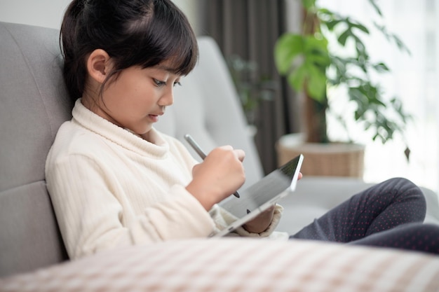 Kleines Mädchen mit Tablet-Spiel im Internet Kind sitzt auf dem Sofa und beobachtet oder spricht online mit einem Freund Kind, das sich morgens im Wohnzimmer entspannt Kinder mit neuem Technologiekonzept
