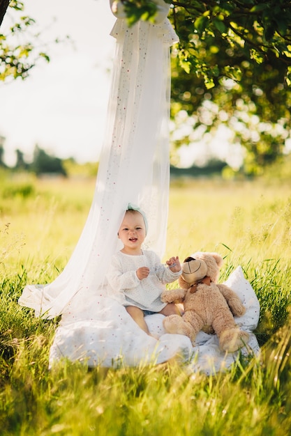 Kleines Mädchen mit Teddybär sitzt auf weißer Decke im sonnigen Sommerpark.