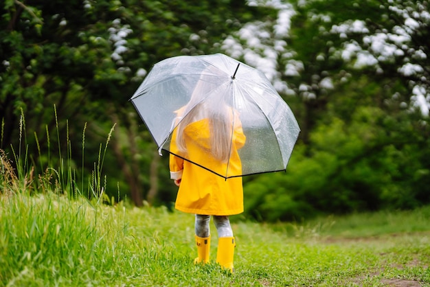 Kleines Mädchen mit transparentem Regenschirm, das im Regen an sonnigen Tagen spielt Schönes Mädchen in einem gelben Mantel.