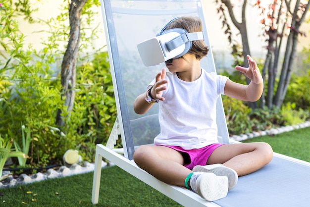 Kleines Mädchen mit Virtual-Reality-Brille, das nach links schaut und auf einer Sonnenliege im Garten ihres Hauses sitzt Metaverse VR-Spiel digital und Simulationskonzept