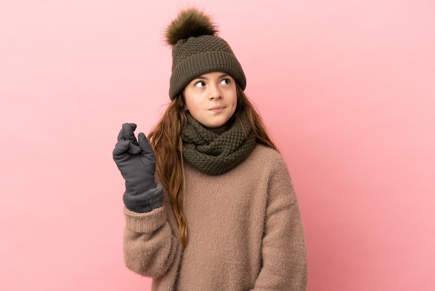 Kleines Mädchen mit Wintermütze auf rosafarbenem Hintergrund mit gekreuzten Fingern isoliert und wünscht das Beste