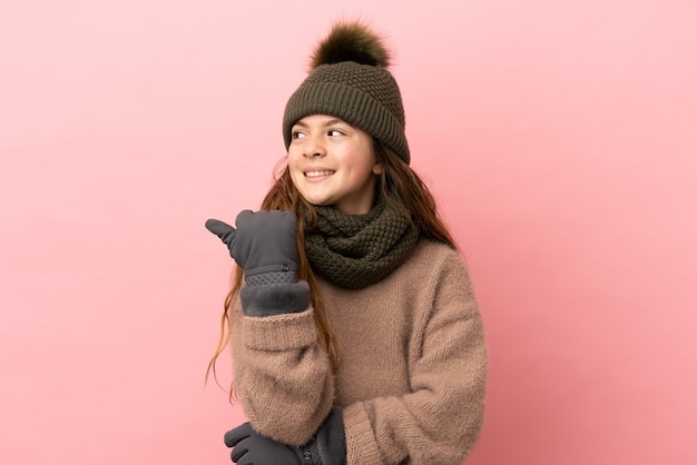 Kleines Mädchen mit Wintermütze isoliert auf rosa Hintergrund, das auf die Seite zeigt, um ein Produkt zu präsentieren