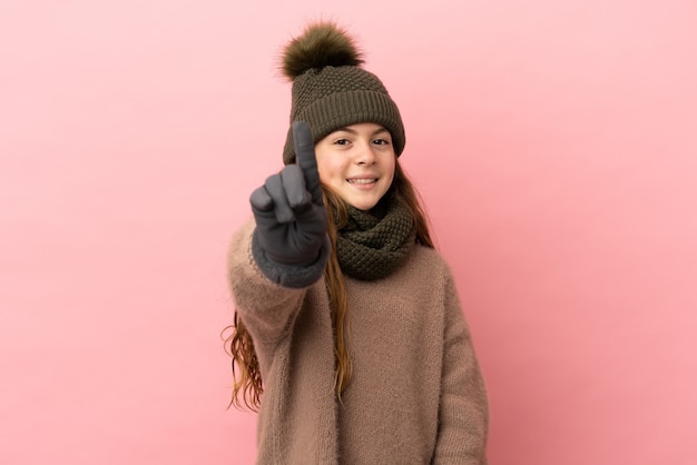 Kleines Mädchen mit Wintermütze isoliert auf rosa Hintergrund, das einen Finger zeigt und hebt