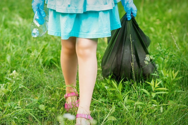 Foto kleines mädchen mit zerknitterter plastikflasche und müllsack in ihren händen beim reinigen des mülls im park