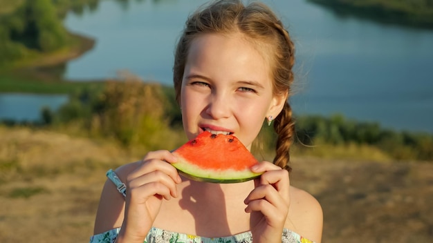 Kleines Mädchen mit Zopf beißt saftige Wassermelone am Flussufer