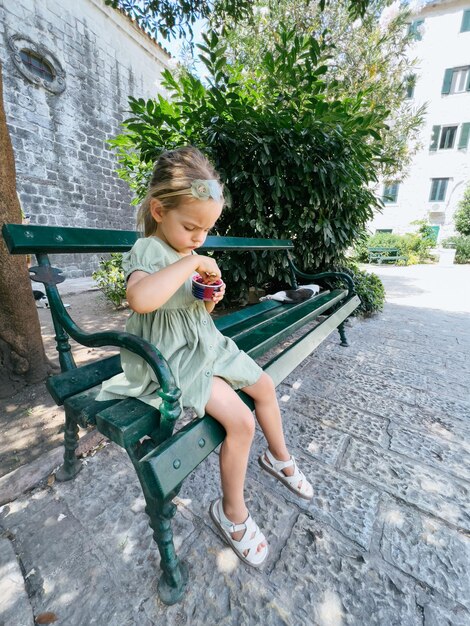 Kleines Mädchen pflückt mit einem Spatel Eis in einer Tasse, während es auf einer Bank sitzt