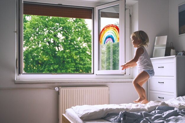 Kleines Mädchen schaut im Frühling aus dem Fenster Nettes Kind auf dem Hintergrund der Malerei Regenbogen am Fenster