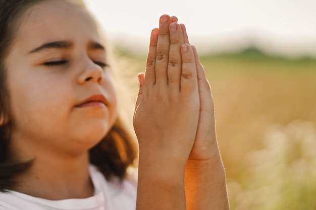 Kleines Mädchen schloss die Augen und betete in einem Feldweizen. Hände im Gebet gefaltet. Religionskonzept