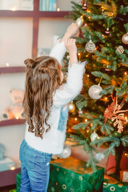 Kleines Mädchen schmückt an Heiligabend einen Baum in einem weißen Pullover