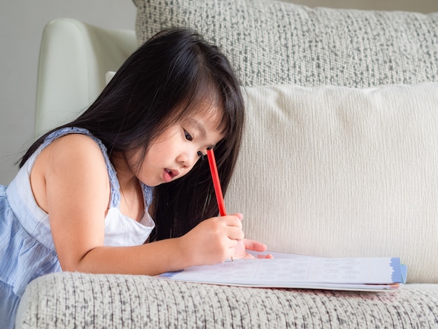 Kleines Mädchen schreibt das Buch mit rotem Bleistift auf das Sofa.