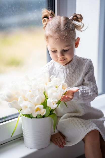 Kleines Mädchen sitzt am Fenster mit Tulpenblumenstrauß Glückliches Kind drinnen Mother39s Day valentine39s Day oder Geburtstagskonzept