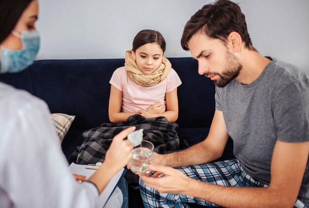 Kleines Mädchen sitzt auf dem Sofa. Ihre Beine und ihr Hals sind bedeckt. Sie sieht aus, wie der Arzt Pulvermedizin in ein Glas Wasser gießt. Junge Mann helfen. Sie sehen konzentriert aus.