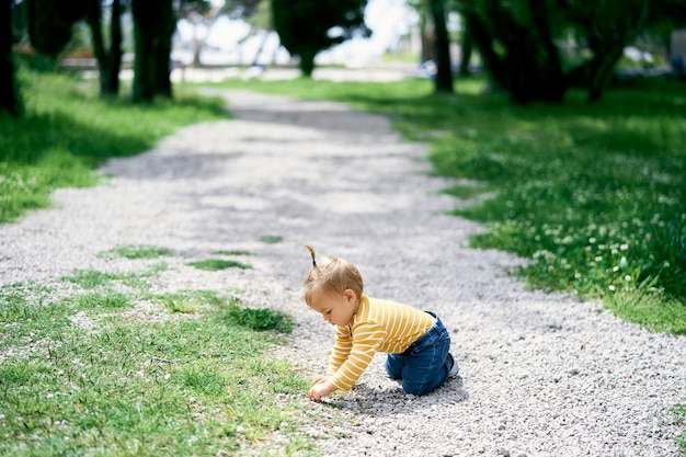 Kleines Mädchen sitzt auf den Knien auf einem Kiesweg in einem grünen Park