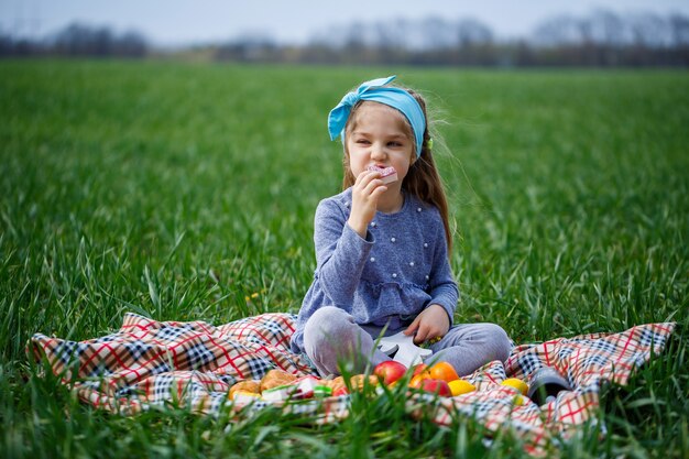 Kleines Mädchen sitzt auf der Tagesdecke und isst Kekse und Marmelade, grünes Gras auf dem Feld, sonniges Frühlingswetter, Lächeln und Freude des Kindes, blauer Himmel mit Wolken