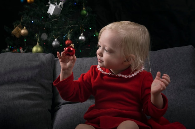 Kleines Mädchen sitzt auf einem Sofa vor dem Hintergrund eines Weihnachtsbaums und betrachtet ein Weihnachtsbaumspielzeug. Neujahr