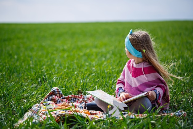 Kleines Mädchen sitzt auf einer Tagesdecke und liest ein Buch mit einem Märchen, grünes Gras auf dem Feld, sonniges Frühlingswetter, Lächeln und Freude des Kindes, blauer Himmel mit Wolken