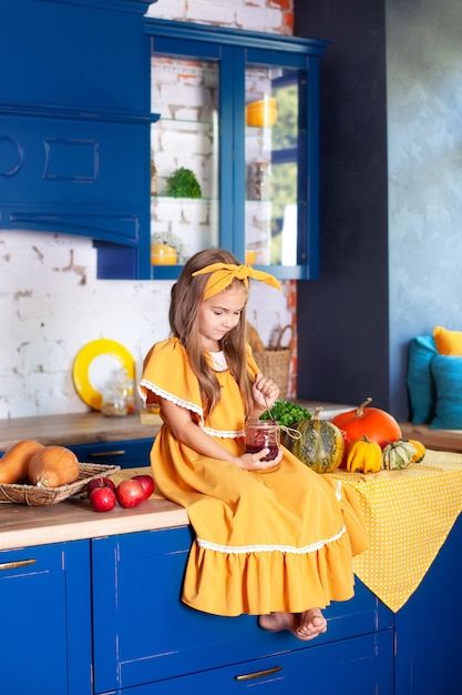 Kleines Mädchen sitzt auf Küchentisch mit Glas Marmelade. Fröhliches Kind in der Küche. Kleiner Koch. Küchenausstattung mit Kürbissen, Äpfeln. Gesundes Essen. Ernte. Herbstdekor. Rustikal.