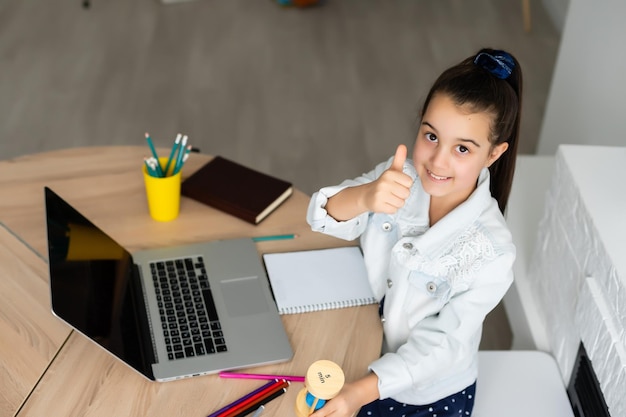 kleines Mädchen sitzt mit einem Laptop am Tisch und hält eine Sanduhr und wartet auf das Ende des Online-Unterrichts