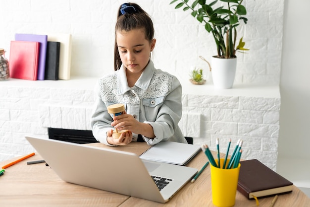 kleines Mädchen sitzt mit einem Laptop am Tisch und hält eine Sanduhr und wartet auf das Ende des Online-Unterrichts