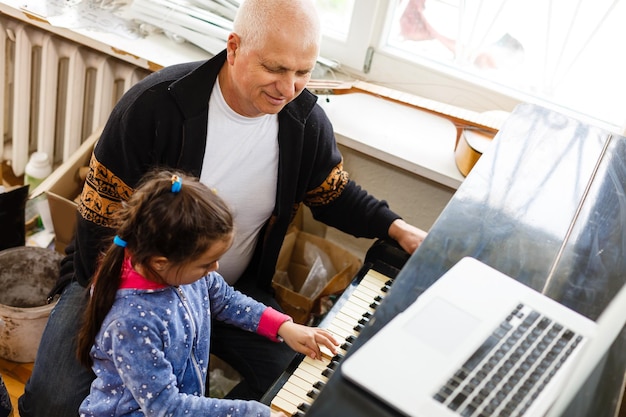 kleines Mädchen spielt zusammen mit Großvater Klavier und lernt online auf einem Laptop