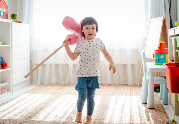 Kleines Mädchen springt und rennt auf Pferdestock im Kinderzimmer.