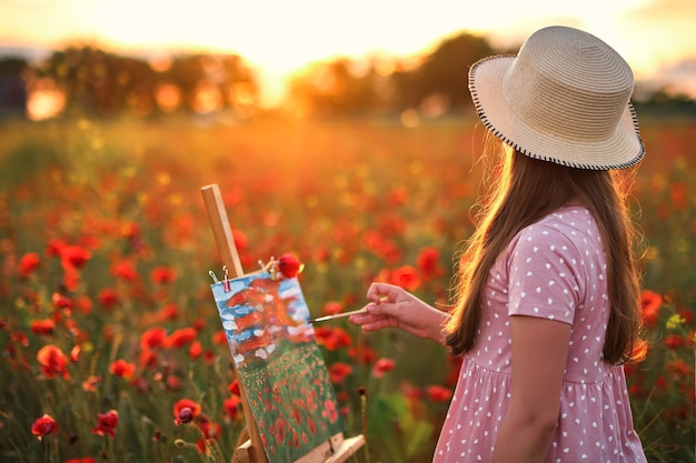 Kleines Mädchen steht im Feld der roten Mohnblumen und malt auf der Leinwand auf einem Zeichenständer a