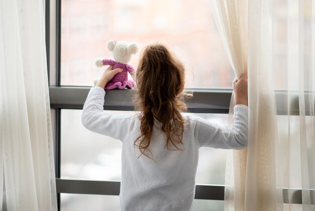 Kleines Mädchen steht in der Nähe eines Fensters mit Teddybär in ihren Händen Lifestile der Kindheit