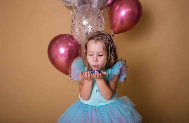 Kleines Mädchen steht mit einem Haufen Luftballons und spielt mit Konfetti auf beigem Hintergrund mit einem Platz für Text