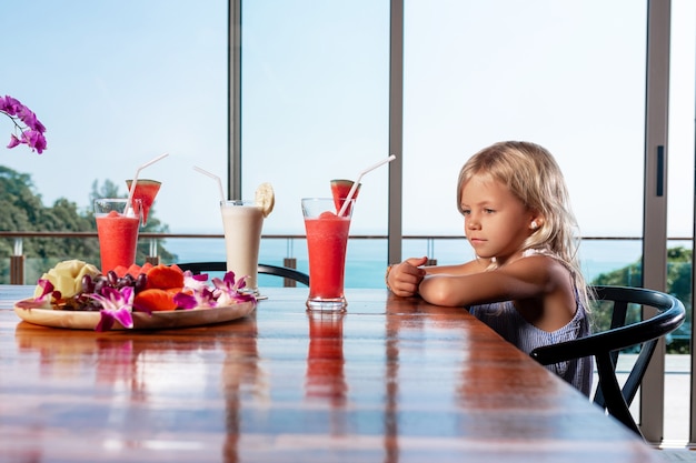 Kleines Mädchen, süßes Mädchen, das am Küchentisch sitzt und tropische Cocktails aus frischen Wassermelonen und Bananen betrachtet