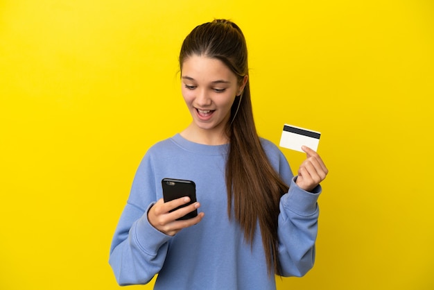 Kleines Mädchen über isoliertem gelbem Hintergrund, das mit dem Handy mit einer Kreditkarte kauft