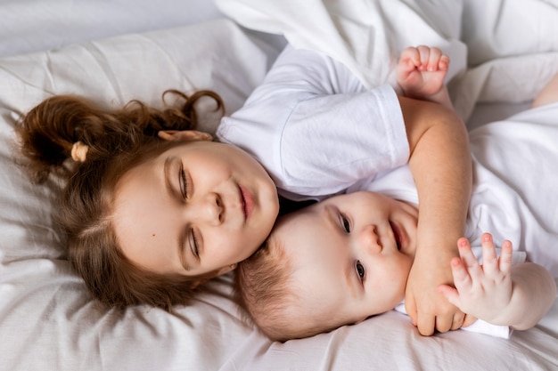 Kleines Mädchen und Baby in weißen Kleidern liegen im Bett auf Bettwäsche Bruder und Schwester sonnen sich im Bett