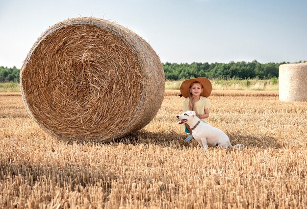 Kleines Mädchen und Hund, die sich an einem Sommertag auf einem Weizenfeld amüsieren. Kind, das während der Erntezeit am Heuballenfeld spielt.