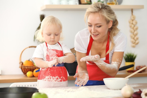 Kleines Mädchen und ihre blonde Mutter in roten Schürzen spielen und lachen, während sie den Teig in der Küche kneten. Hausgemachtes Gebäck für Brot, Pizza oder Plätzchen backen. Familienspaß und Kochkonzept.