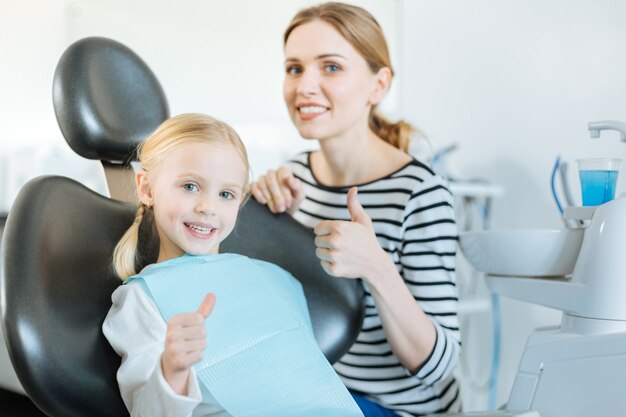 kleines Mädchen und ihre geliebte Mutter zeigen Daumen hoch, während das Mädchen auf dem Zahnarztstuhl sitzt und auf die Behandlung wartet