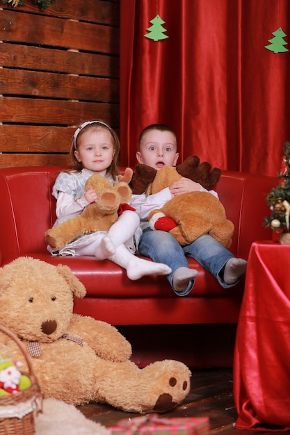 Foto kleines mädchen und junge auf sofa im weihnachtsstudio in den roten farben. weihnachtsbaum und teddybär an der wand.