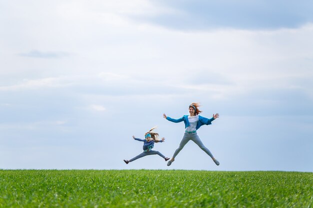 Kleines Mädchen und Mutterfrau laufen und springen, grünes Gras auf dem Feld, sonniges Frühlingswetter, Lächeln und Freude des Kindes, blauer Himmel mit Wolken