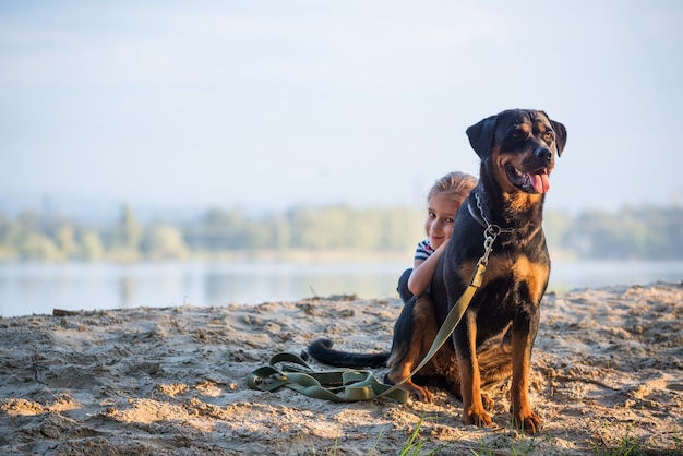 Kleines Mädchen und Rottweiler Hund kuscheln am Rand einer Klippe und genießen einen warmen Morgen im Wald