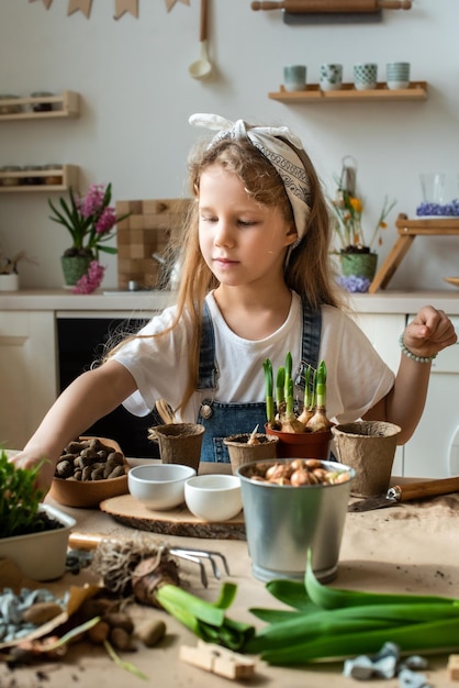 Kleines Mädchen verpflanzt Blumen und Zimmerpflanzen, ein Kind in einem Bandana pflanzt Blumenzwiebeln und Microgreens