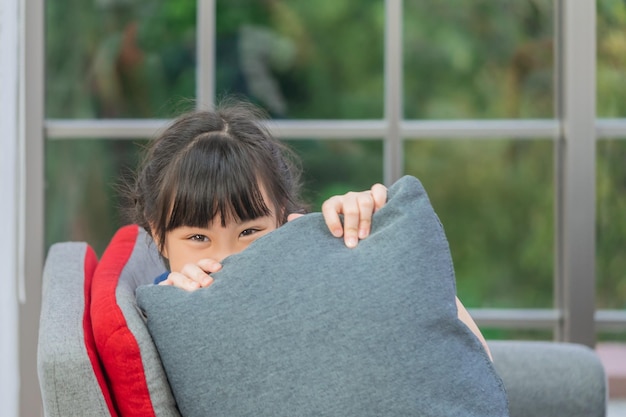 Foto kleines mädchen versteckt sich hinter kissen mit lächeln und verspieltem gesicht auf einem grauen sofa im wohnzimmer