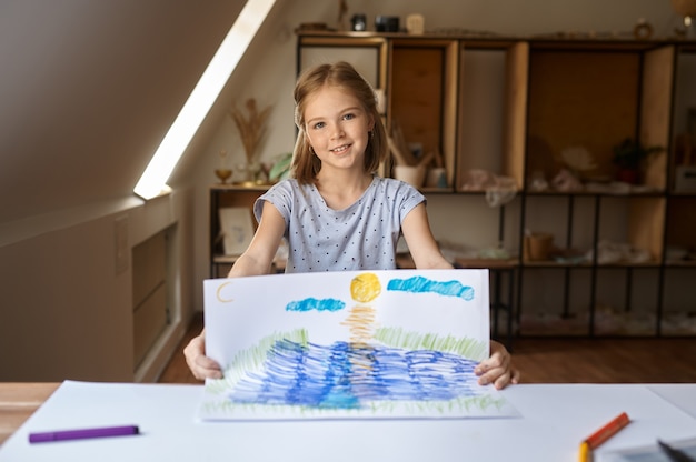 Kleines Mädchen zeigt ihre Zeichnung, Kind in der Werkstatt. Unterricht an der Kunstschule. Junger Maler, angenehmes Hobby, glückliche Kindheit. Kreative Entwicklung