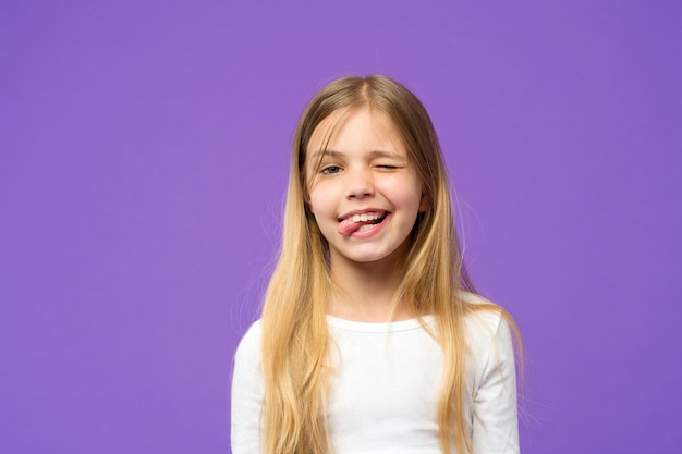 Kleines Mädchen zeigt Zunge. Kind mit lustigem Gesicht auf violettem Hintergrund. Nettes Kind mit Schönheitsblick und langen blonden Haaren. Beauty-Look und Haarpflege. Fröhliche Kindheit und Kinderbetreuung, Kopierraum.